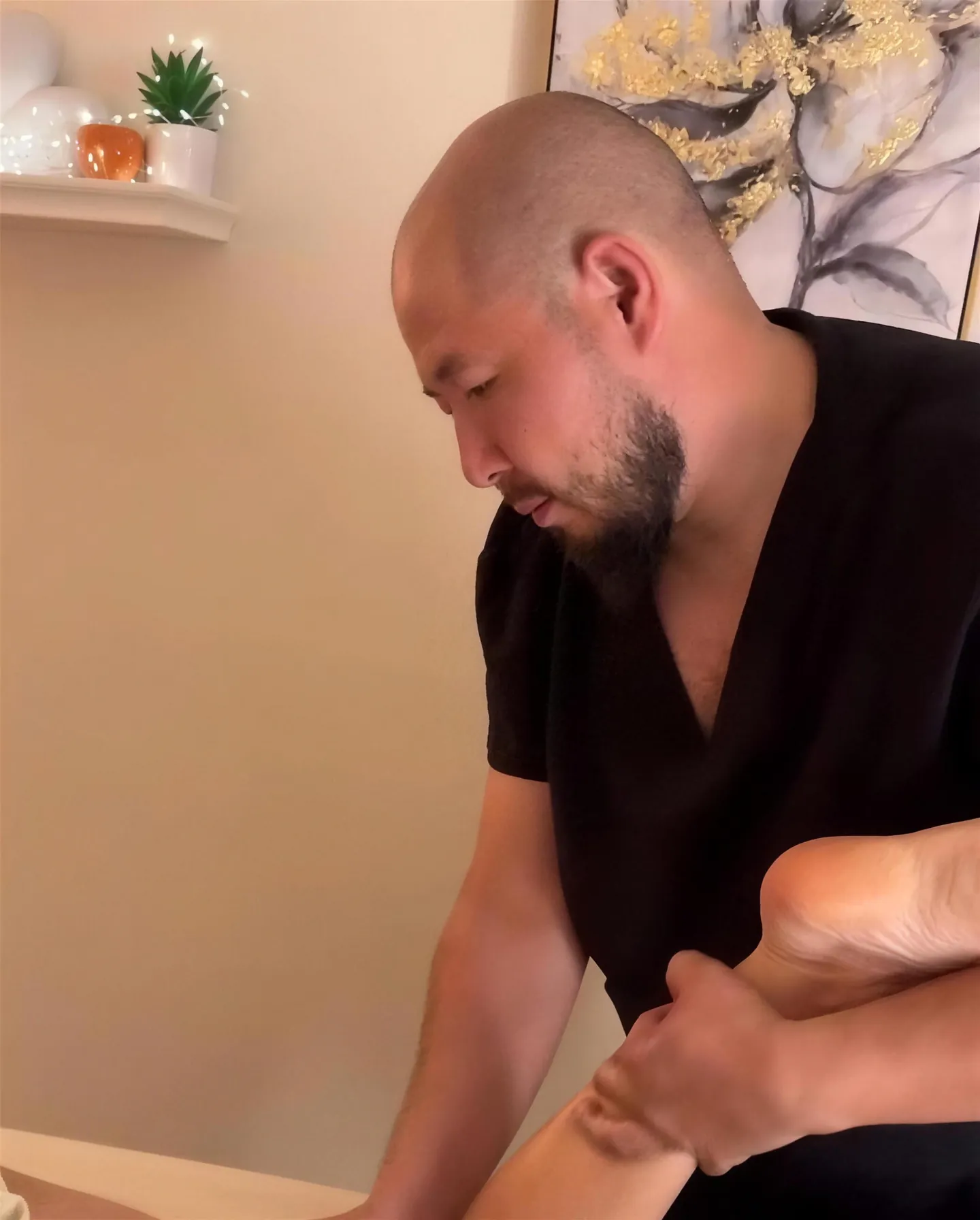 Tony, focused and calm, working on a client's foot, with a framed painting and a shelf behind him